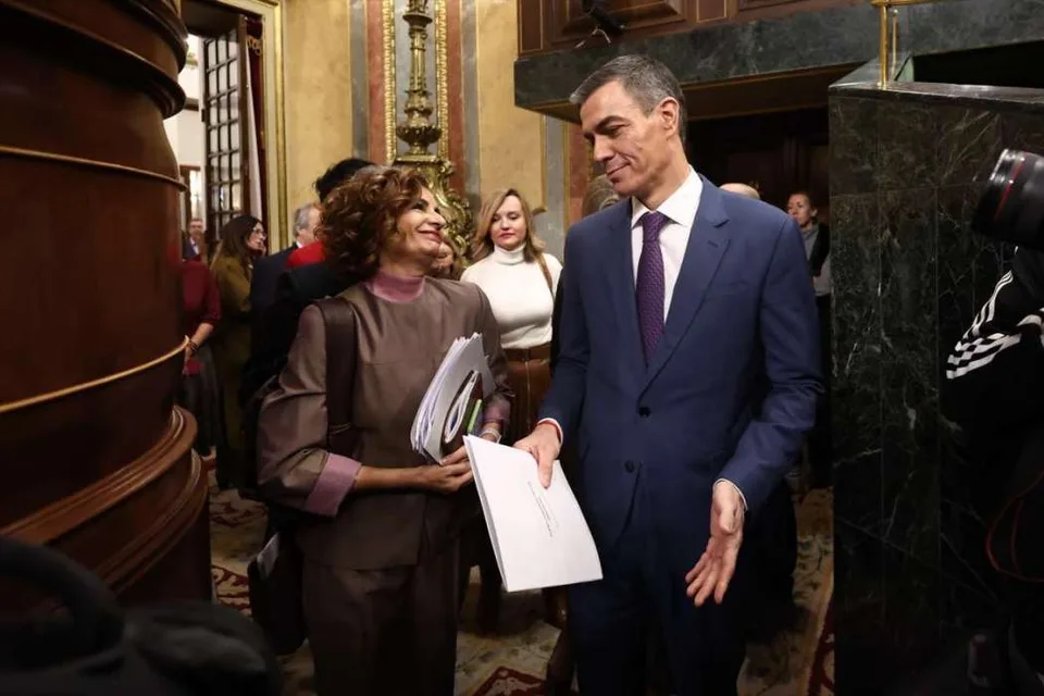 Dos dirigentes políticos conversan junto a otros asistentes en un salón institucional, con documentos en la mano.